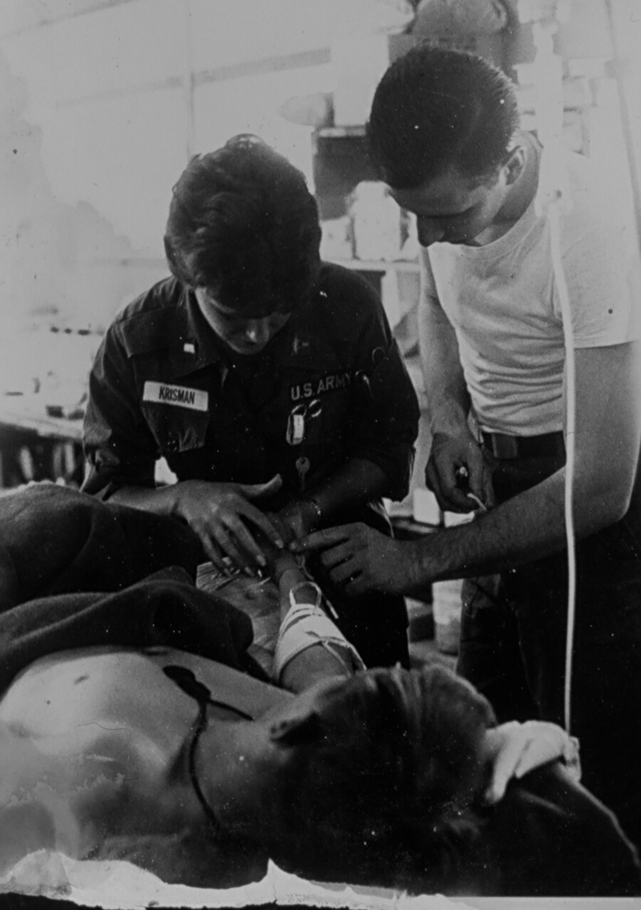 A nurse and doctor standing over a patient. The nurse is wearing a nametag that reads "Krisman."