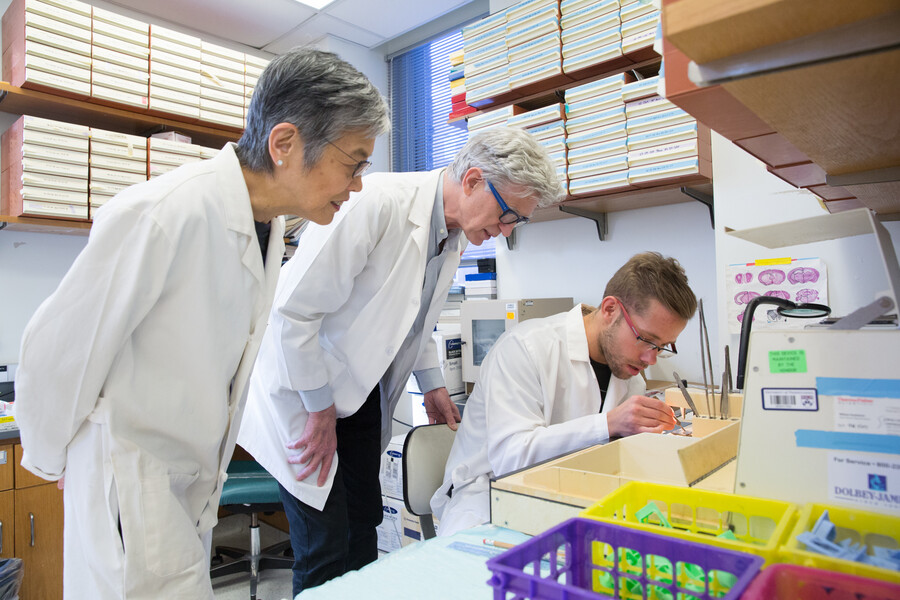 Virginia Lee and John Trojanowski oversee a student in a lab, seated. 