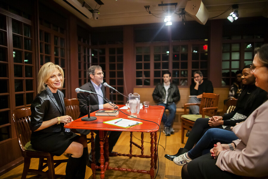 Two people sitting at a wooden table with microphones look out at a crowd.