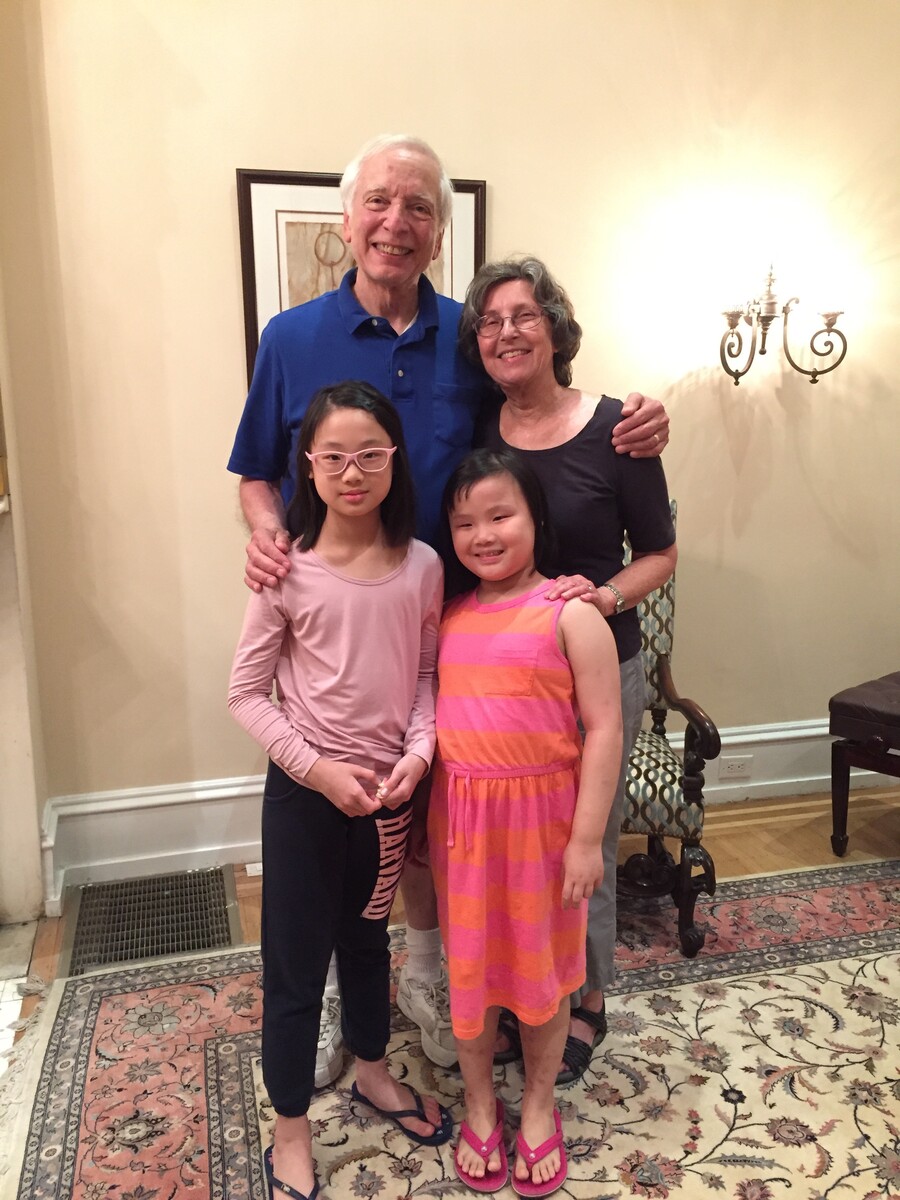 Four people—an older couple and two children—standing on a floral run in front of a pictures hanging on the wall and a light sconce.