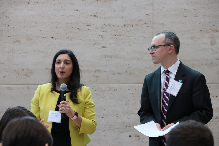 One person in yellow jacket speaks into a microphone, another person stands next to them, holding a piece of paper