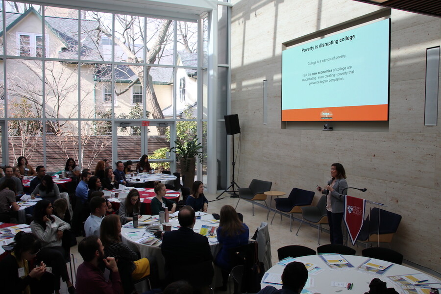 Person speaks standing in front of room full of people seated at round tables, underneath a large screen which says "Poverty is disrupting college."