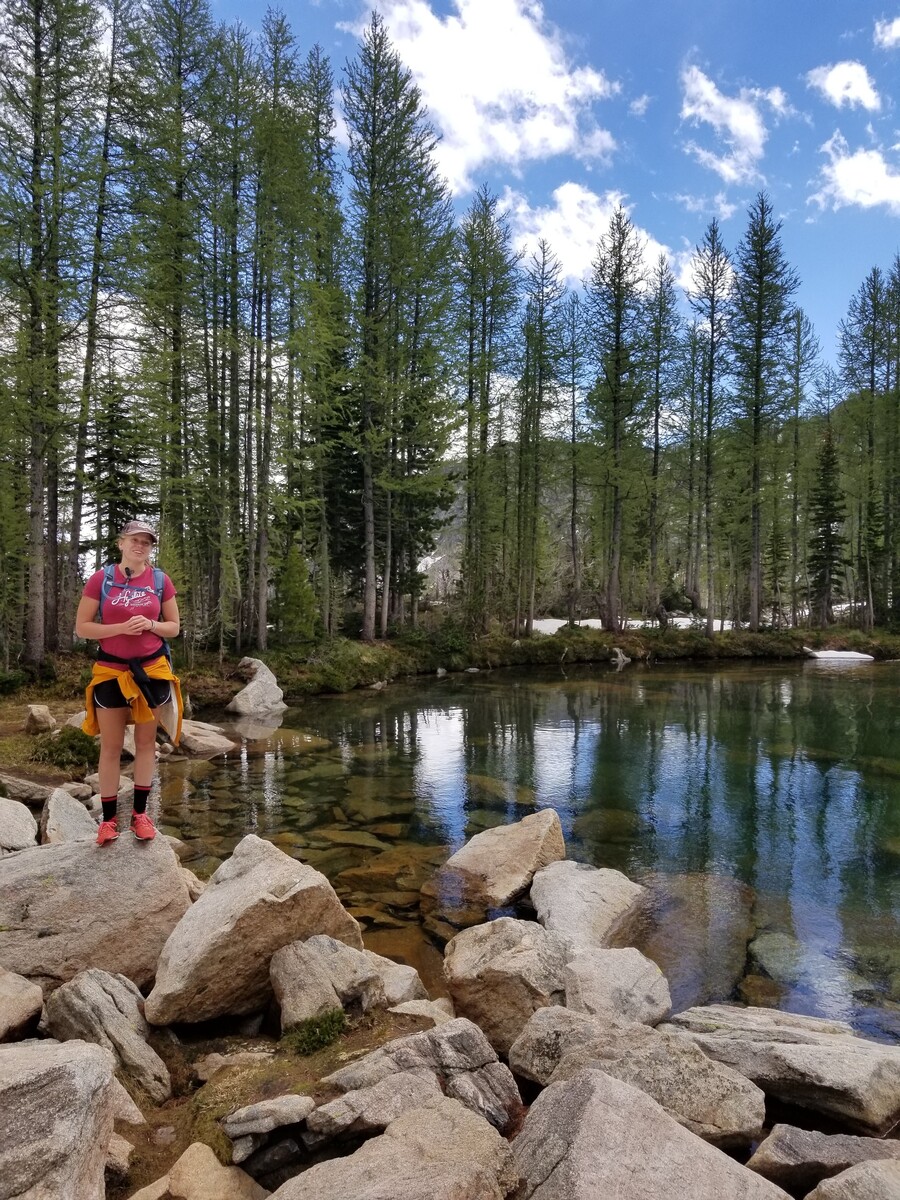 A person in athletic gear and a baseball cap in nature, standing on rocks in front of water and trees.