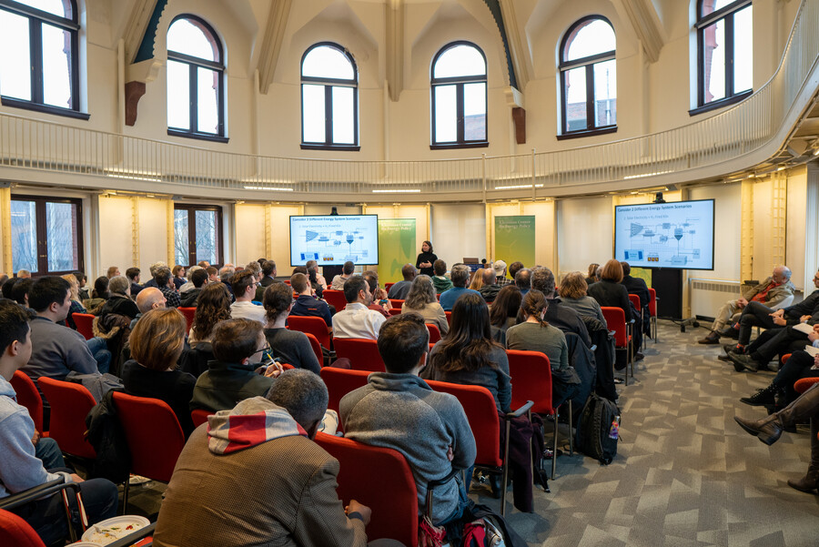 A crowd in red chairs listening to a speaker in front of the room between two projectors.