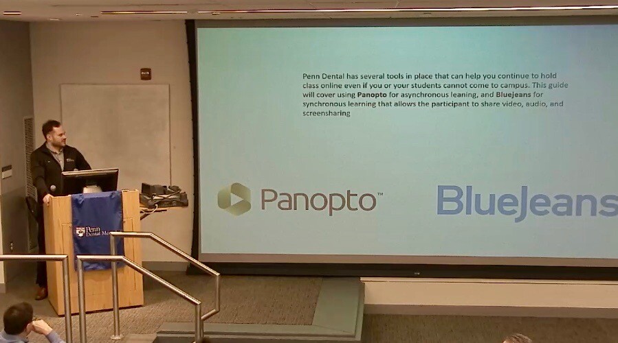 Person standing at podium with computer next to a large screen in a workshop for faculty on remote teaching.