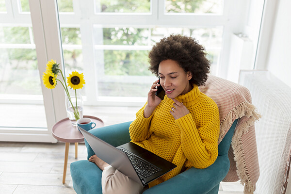 person seated in a chair at home with a laptop on their lap, holding a smartphone to their ear, with a side table holding a cup of tea and sunflowers and bright sunlit windows behind them