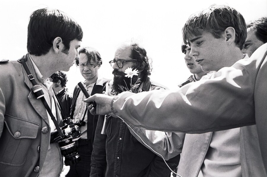 Man with beard and glasses holds a daisy while being interviewed by student reporters in 1970