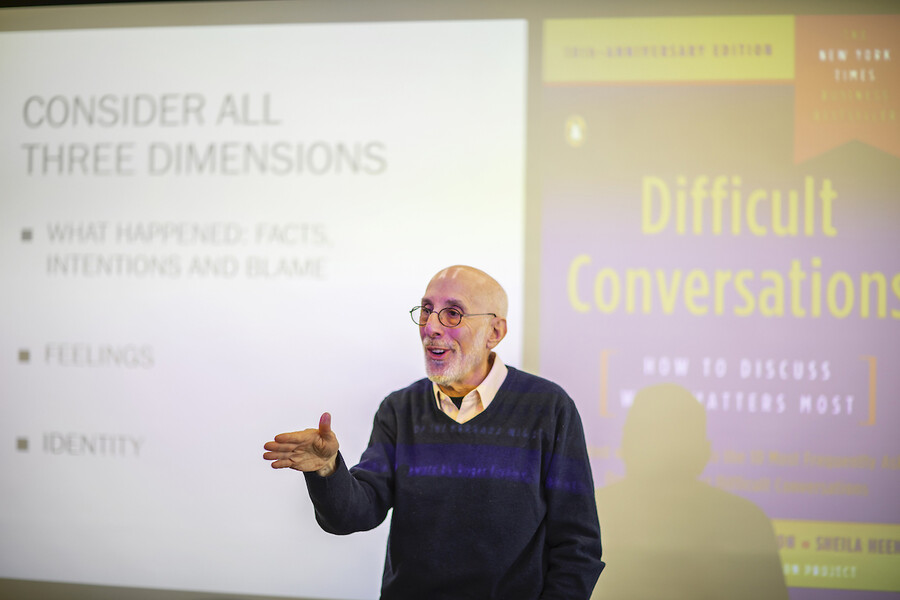 Person gestures with right hand as stands in front of screen that reads "difficult conversations"