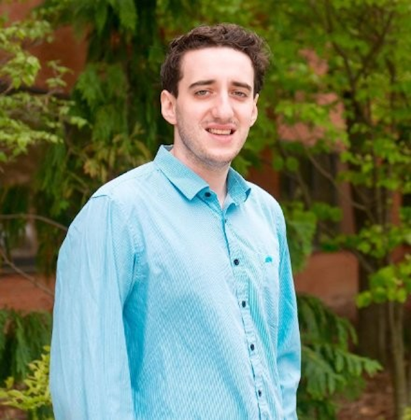 Man in blue shirt standing in front of brick buildings and green trees smiles at the camera