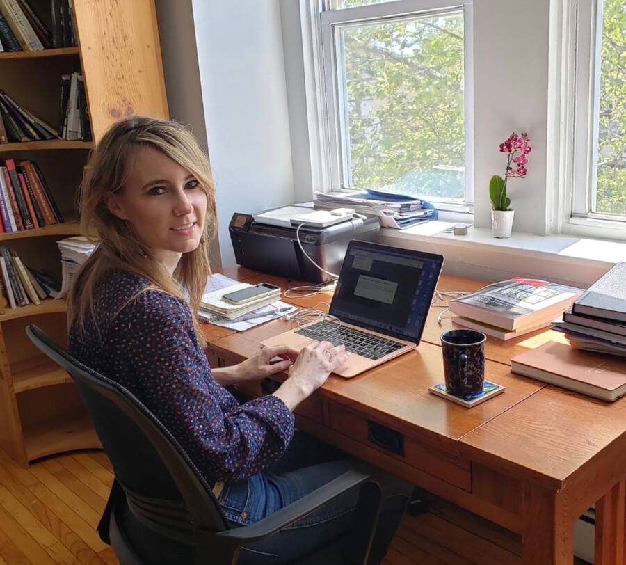 A woman sits at a desk, working on a laptop in front of two windows