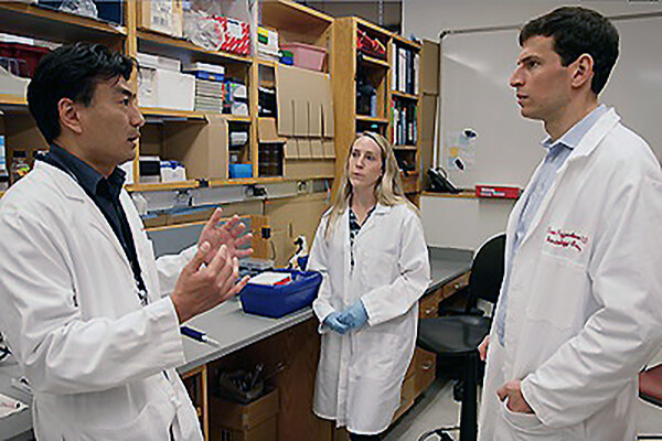 &nbsp;Taku Kambayashi, Ruth-Anne Langan Pai, and David Fajgenbaum stand talking in a lab.