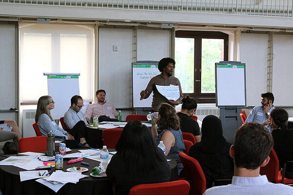 Person standing in front of a group of people seated around tables presenting talking points on paper.