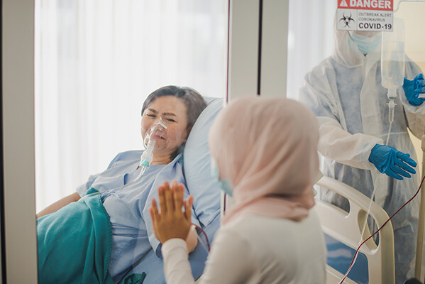 Person in hospital bed with COVID-19 presses their hand against a glass window with a loved one on the other side.