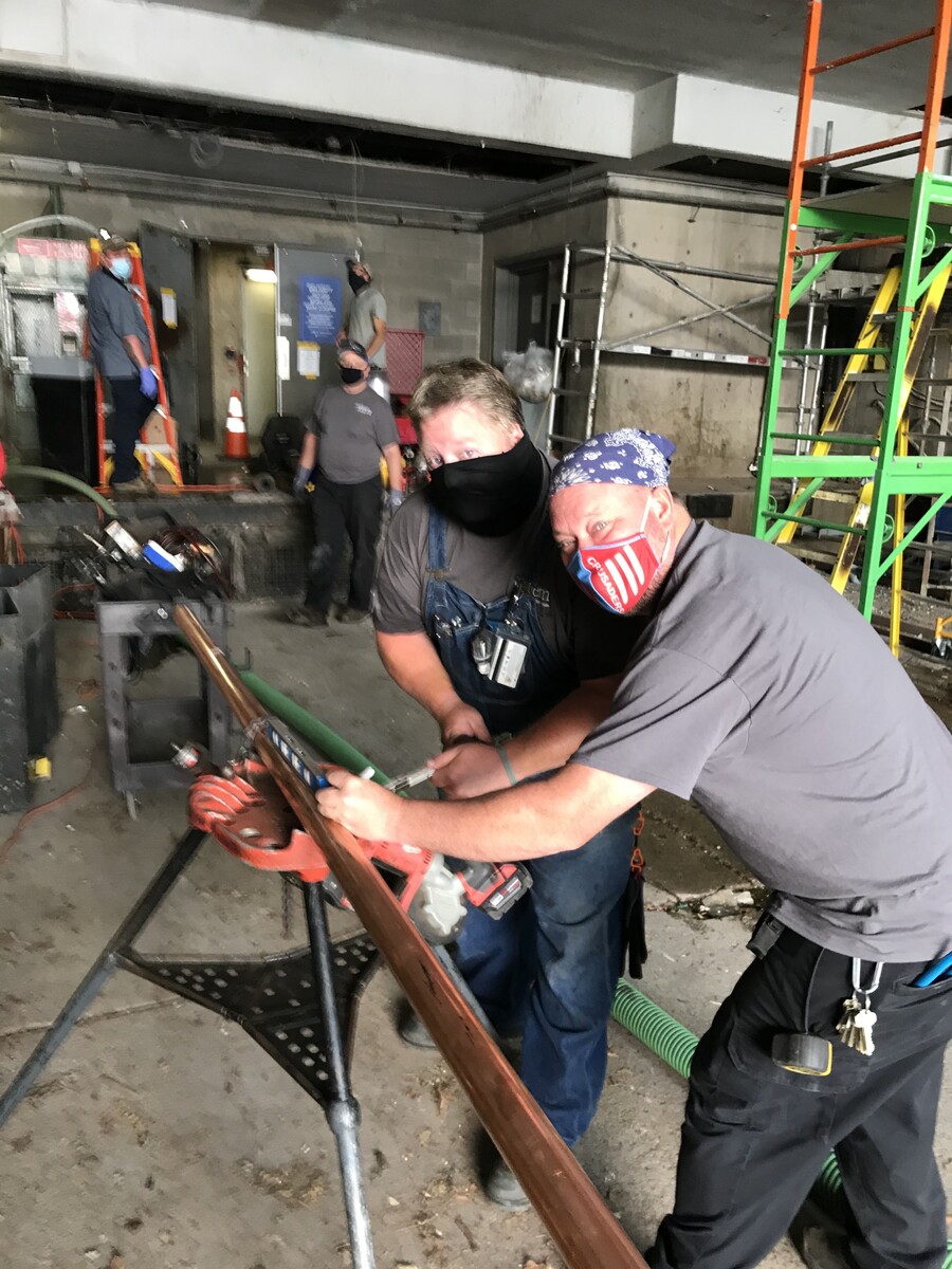 Two people wearing masks use tools in the foreground, with ladders and a masked person in the back 