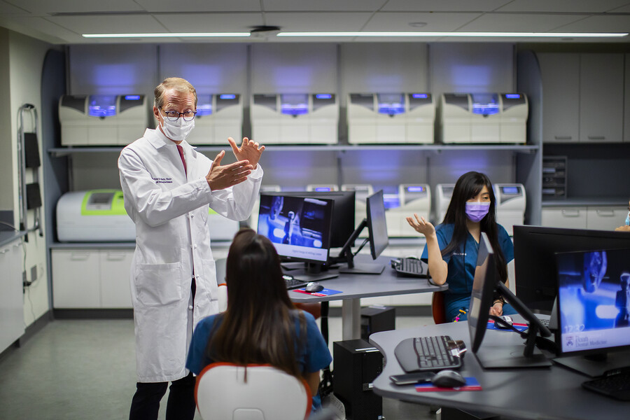 Markus Blatz standing in a dental digital lab wearing a white coat and a face mask, talking to two students seated at tables with computers.