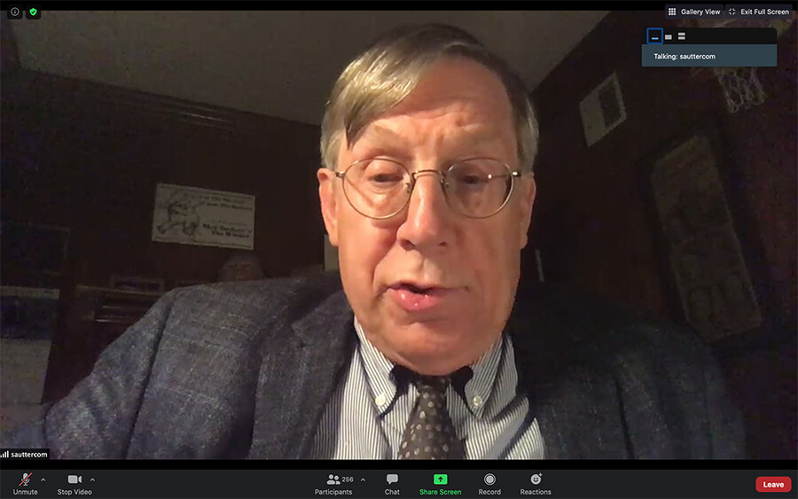 A man with glasses and a grey suit and black tie with dots speaks during a Zoom call