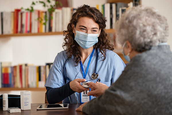A home health aid wearing a face mask uses an oximeter on an elderly patient at home.