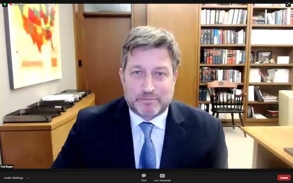 Man sits in office in front of book shelf, looking into camera wearing blue tie and suit jacket