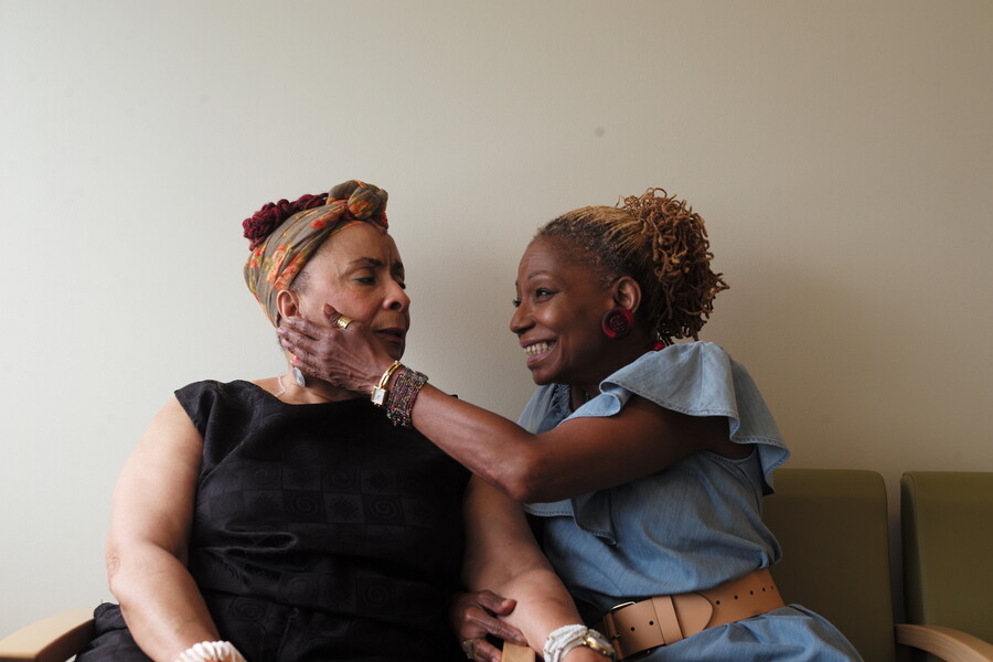 Two people sitting next to each other on a couch holding hands. One is smiling and touching the other's face.