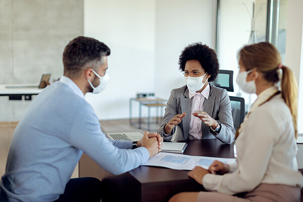 Three masked individuals dressed in business attire sit at a desk, one is African American, discussing plans or a possible partnership or investment.