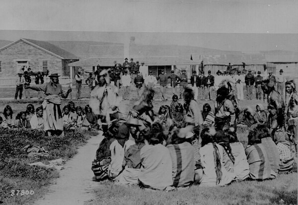 Members of the Shoshone tribe sit on the ground and stand as members of the US military stand behind them and sit on horseback