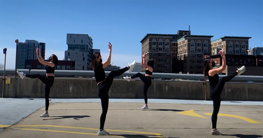 Four dancers wearing masks performing in an outdoor parking area.
