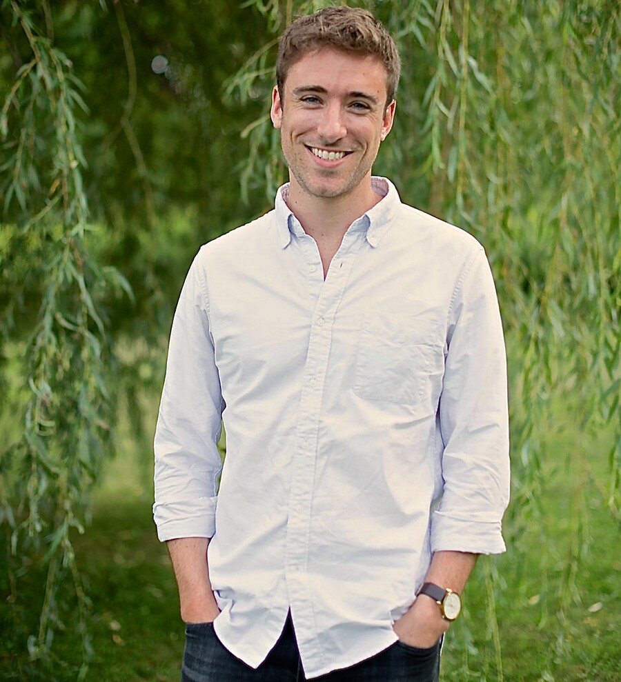 A person standing outside wearing a light-blue button down shirt, with blurred trees in the background.
