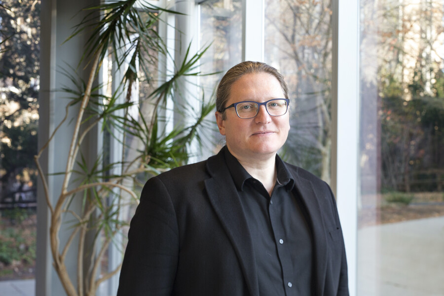 A person in a black shirt and black blazer standing front of tall windows and a spindly plant.