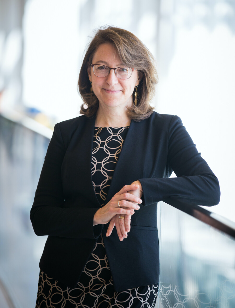 A person leaning against a glass railing, one elbow on the rail, the other hand on top of the leaning hand. The person is wearing a black dress with gold intertwining circles, a black sweater, glasses and gold earrings.