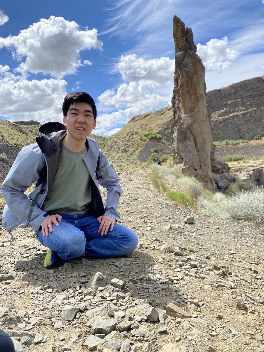 A man sits crossed legged on the dirt as dusty hills covered in green scrub rise behind him with a blue sky and puffy clouds