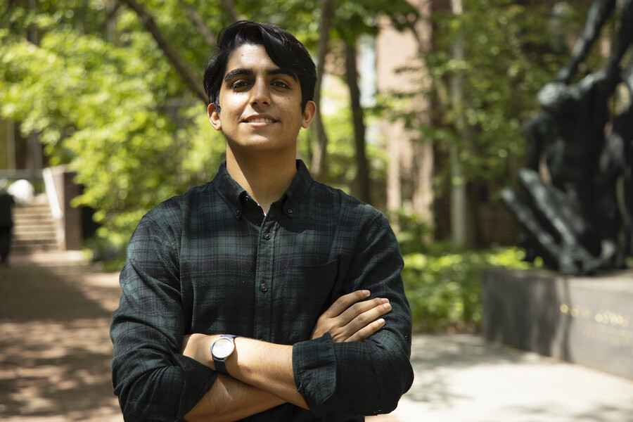 Man stands outside on a brick path smiling at the camera with his arms crossed,  with green trees behind him