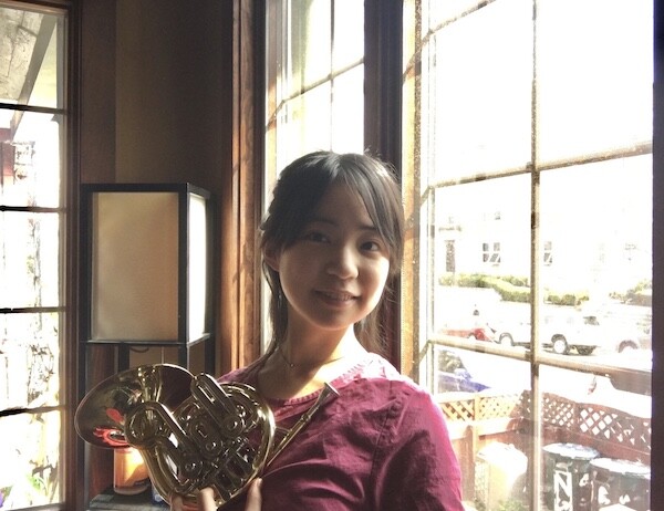 Woman in reddish pink top holds a french horn by a window that looks out onto a street with parked cars
