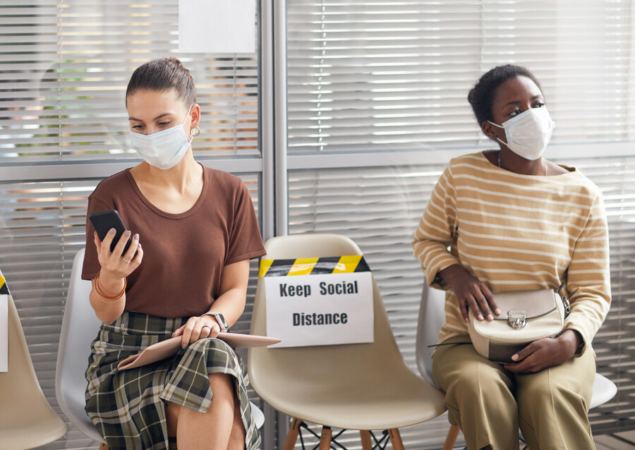 Two people, one Black one white, sitting in a waiting room wearing masks.