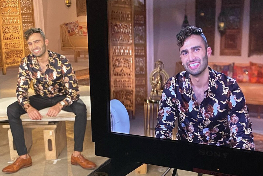 Dillon Patel seated and smiling behind a large television screen filming him.