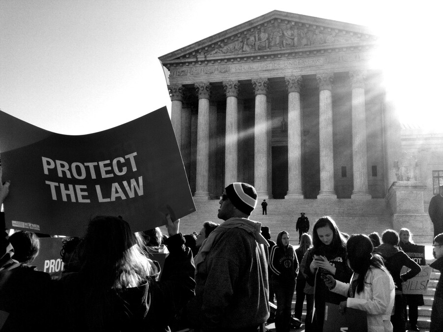 Group of people outside the steps of the Supreme Court, one person holds a large sign that reads PROTECT THE LAW.
