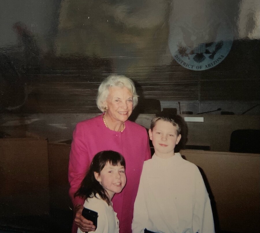 Woman with gray hair in pink suit smiles standing behind two children, a girl on her left and a boy on her right
