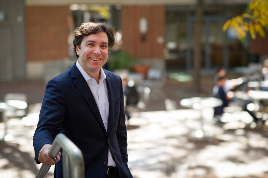 Man in a white shirt and blue blazer holds onto a metal staircase railing with blurry cafe table behind him and a red brick and glass building.