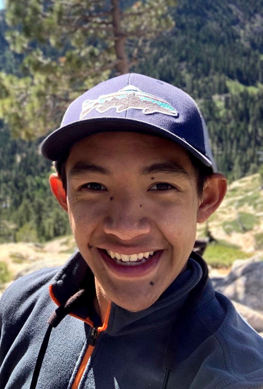 Man in a baseball cap takes smiles into the camera on a sunny day with a forested hillside in the distance behind him