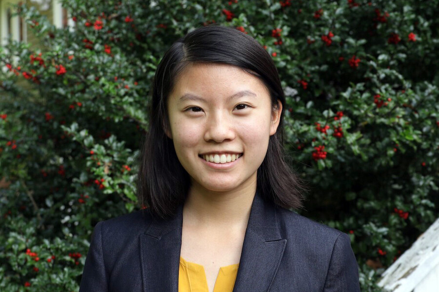 Girl in black blazer and yellow shirt smiles in front of bush covered in red flowers