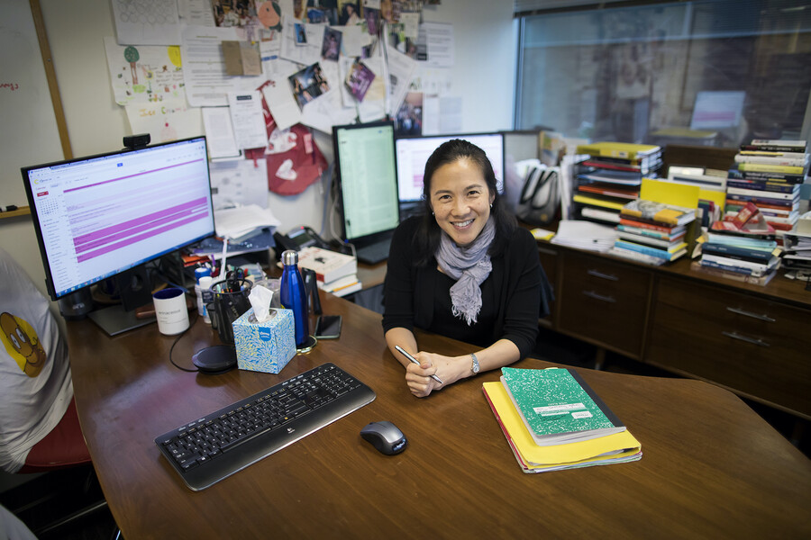 Angela Duckworth at her desk. In the foreground on a brown table sit a composition book and folders, a computer mouse and keyboard, a tissue box, water bottle, and pencil holder, in front of a computer screen. In the background are stacks of books, two additional computer screens, and a wall covered with papers. 