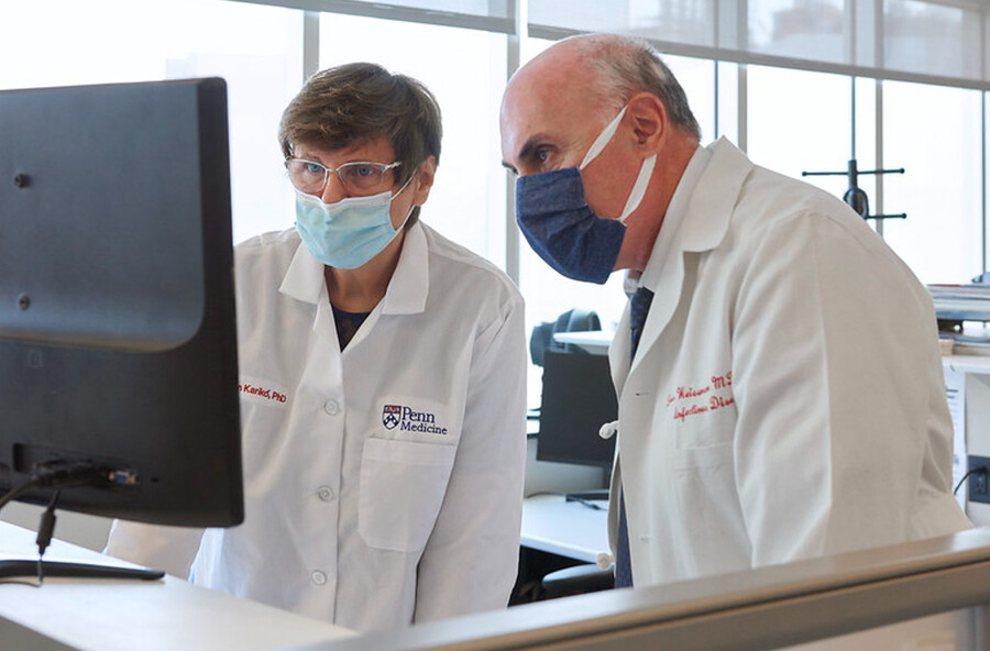 Katalin Karikó and Drew Weissman look at a computer monitor wearing face masks. 