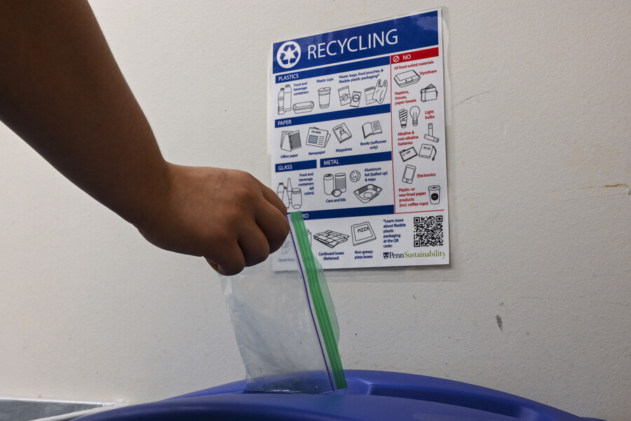 A person's hand disposing of a plastic bag into a blue bin with a Recycling sign above