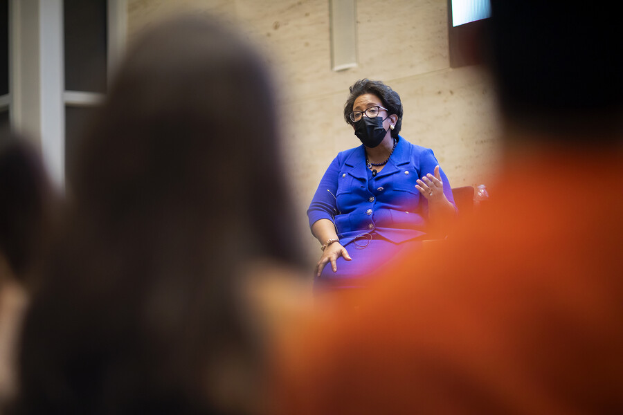 Former attorney general of the United States Loretta Lynch, wearing glasses, a black surgical mask and a blue suit dress, is seen gesturing on a stage from between the blurry backs of two people in the audience