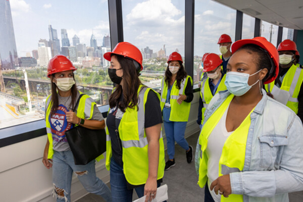 A group of people walking through the Pavilion wearing face masks, hard hats, and safety vests.