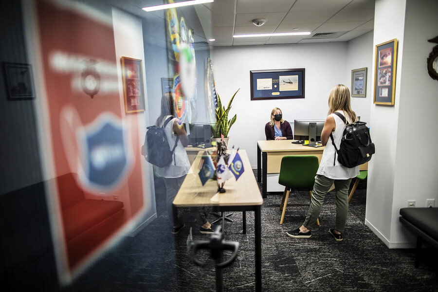A woman wearing a mask sits at a desk in an office, speaking to a student standing up, wearing a backpack