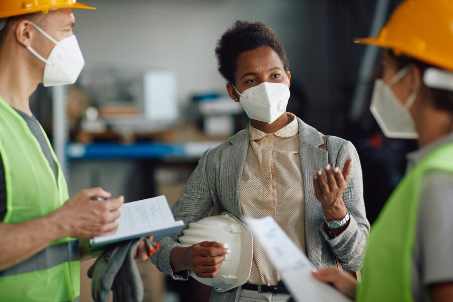 Boss wearing a mask holding a hard hat talks with two employees wearing masks, hard hats, and safety vests.