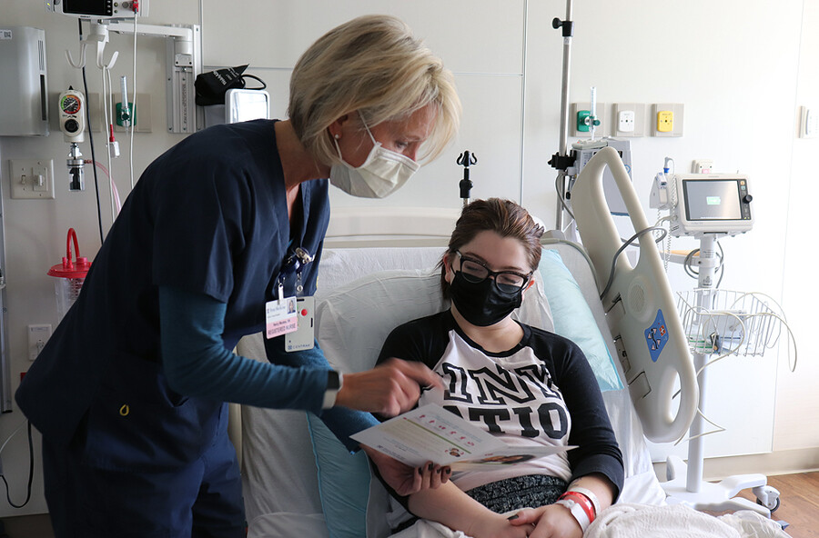 Pregnant person lying in a hospital bed wearing a face mask looking over a sheet of paper a doctor is showing them.