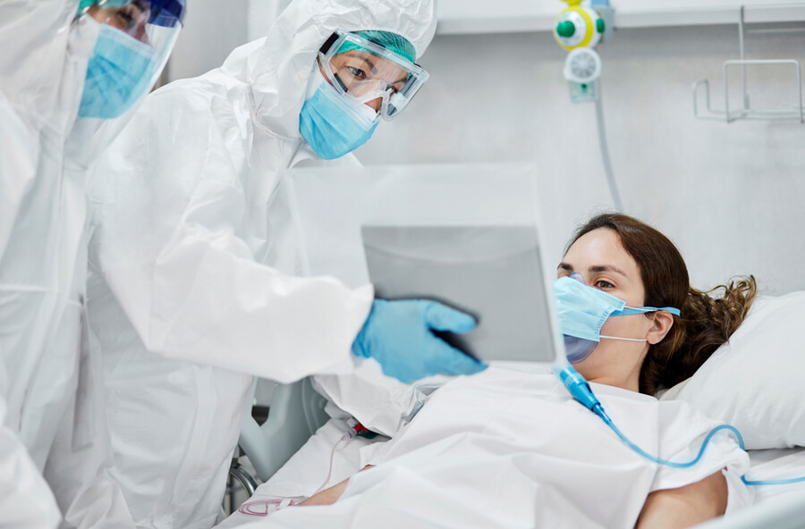 COVID-19 patient in a hospital bed wearing a mask with two medical personnel beside them in full PPE.