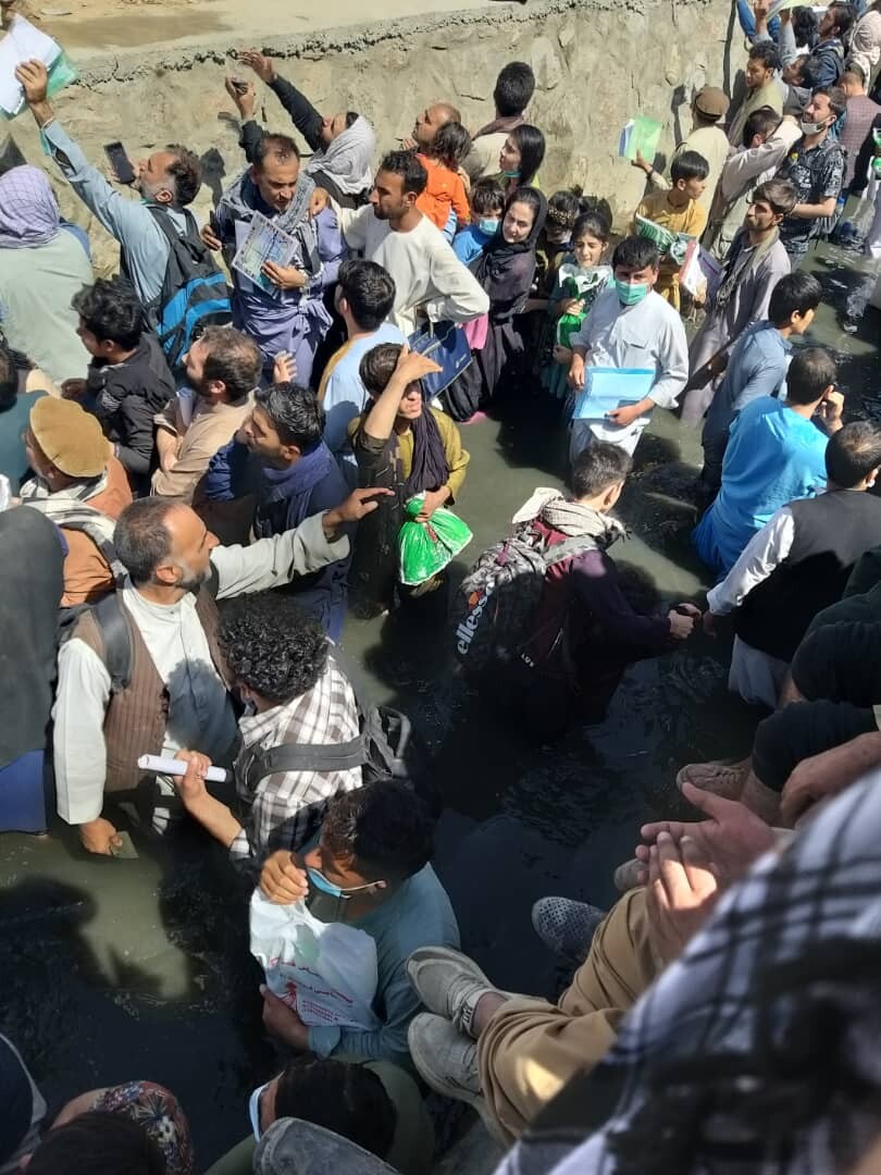 Many people cluttered together in knee-high water, some shielding their eyes, others, holding bags or wearing backpacks.
