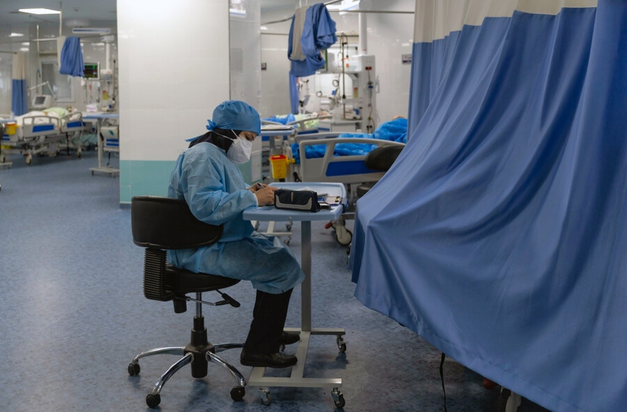 A nurse seated at a work station in a large hospital room with patient beds.